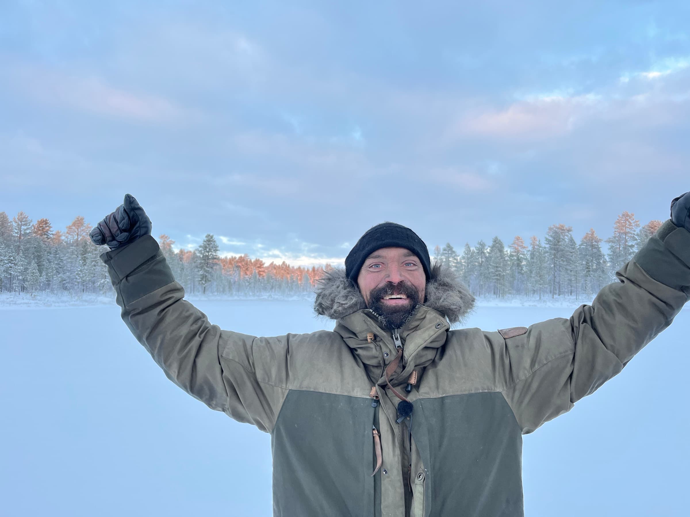 Rasmus Kragh løfter armene og smiler bredt på en frossen, snedækket sø med solbelyst nåleskov bagved